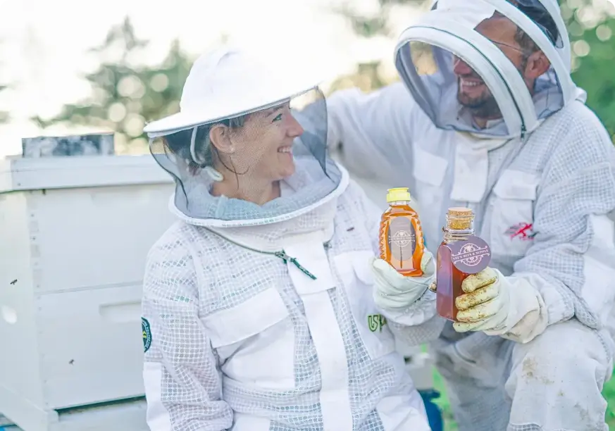 Two people in bee suits holding bottles of honey.